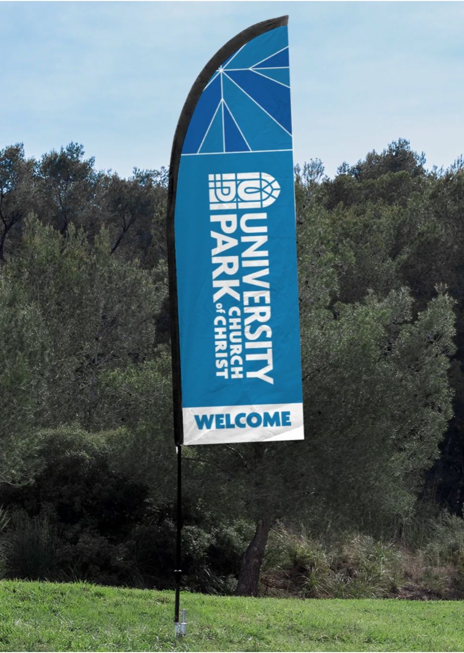 Feather flag staked in grass before a treeline, carrying the stacked logo above the word Welcome.